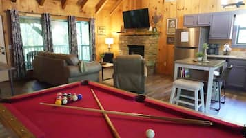 A cozy room with a pool table, a fireplace, a television, and a kitchen area.