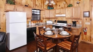 A wooden cabin-style kitchen with a white refrigerator, stove, and dishwasher. There is a dining table set with plates and glasses. - Moonshine Holler (Sevierville)