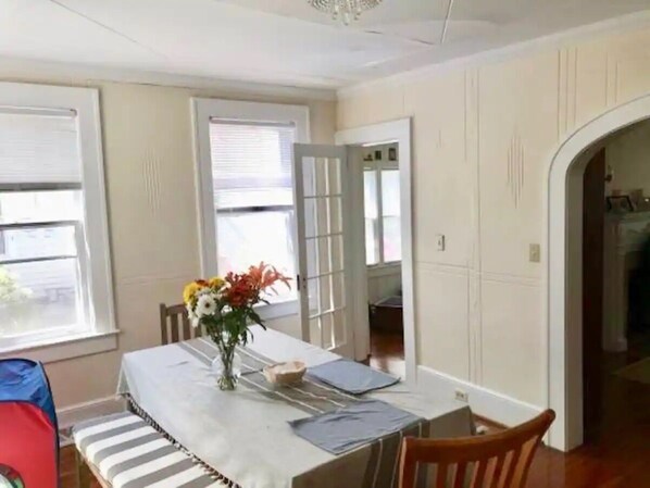 A dining area with a table set for two, a vase with flowers, and a view of the kitchen through an open door.