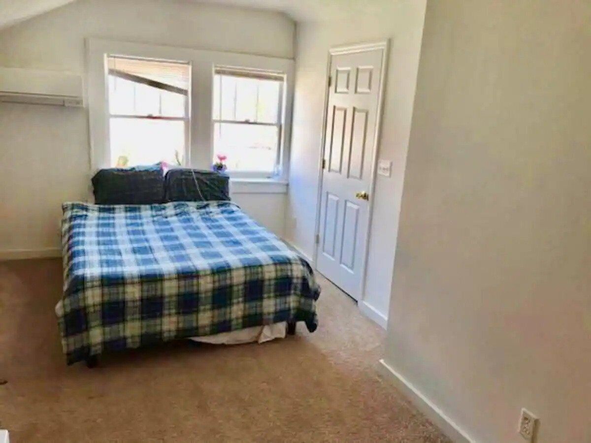 Un dormitorio con cama, dos ventanas, una puerta y piso alfombrado.