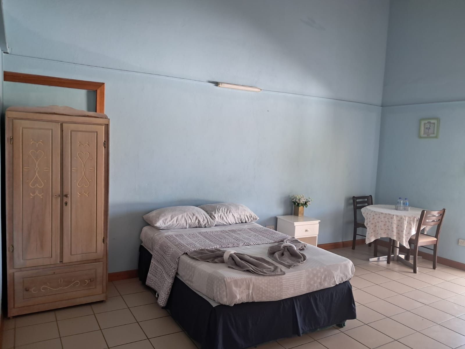 A bedroom with a bed, a wardrobe, a small table with a chair, and a wall-mounted shelf.