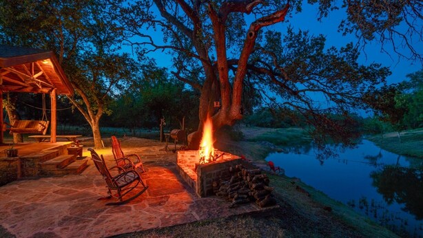 An outdoor seating area with a fire pit, wooden furniture, and a view of a lake at dusk. - Private & Seculded Cabin on Comanche Creek with 73 Acres to Roam (Llano)