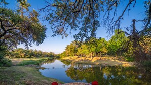 A serene riverside setting with two red chairs and a stone table, surrounded by lush greenery and trees reflecting in the calm water. - Private & Seculded Cabin on Comanche Creek with 73 Acres to Roam (Llano)