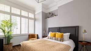 A bedroom with a large bed, a chair, a bedside table, a lamp, a window with blinds, and a potted plant.