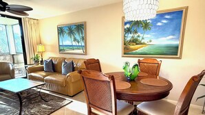 Un salon avec un canapé beige, une table basse, une table à manger avec des chaises et deux tableaux représentant des scènes tropicales.