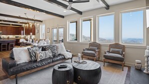 A spacious living area with a leather sofa, two armchairs, a coffee table, and a ceiling fan. A kitchen is visible in the background with wooden cabinets and a dining area.