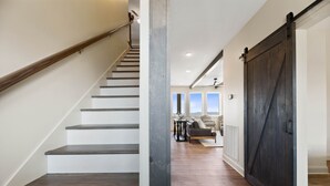 A staircase with wooden steps and a metal handrail, leading to a living area with a large dark wooden door and a view of the outdoors.