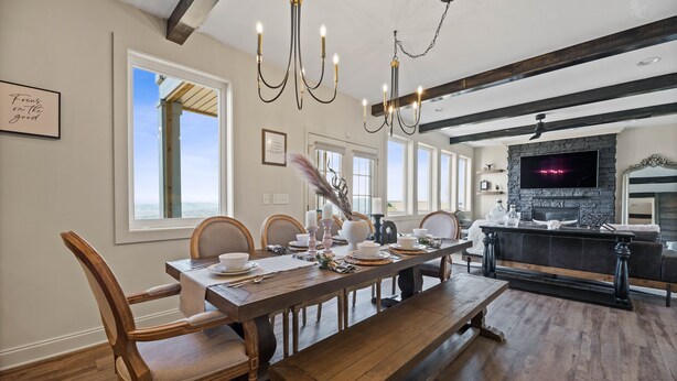 A dining area with a wooden table set for six, surrounded by wooden chairs. A bench is placed in front of the table. The room has a stone fireplace and a television mounted above it.