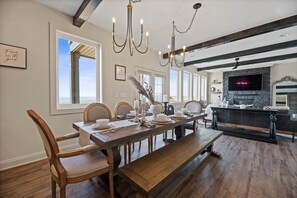 Un comedor con una mesa de madera dispuesta para seis personas, rodeada de sillas de madera. Un banco se encuentra frente a la mesa. La habitación tiene una chimenea de piedra y un televisor montado encima.