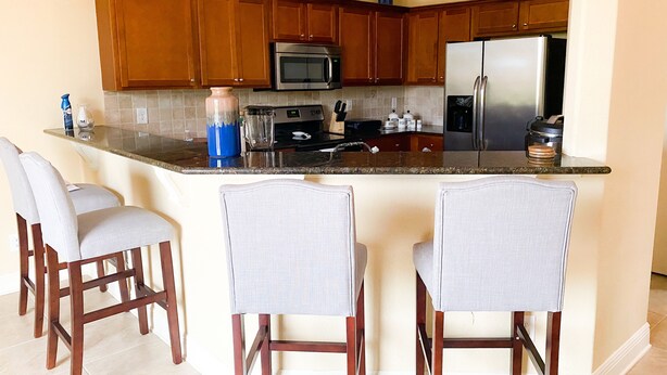 A kitchen with wooden cabinets, a stainless steel refrigerator, a microwave, and a coffee maker. There are three bar stools with cushioned seats. - Cozy home on the water with plenty of sleep space (Fort Myers)