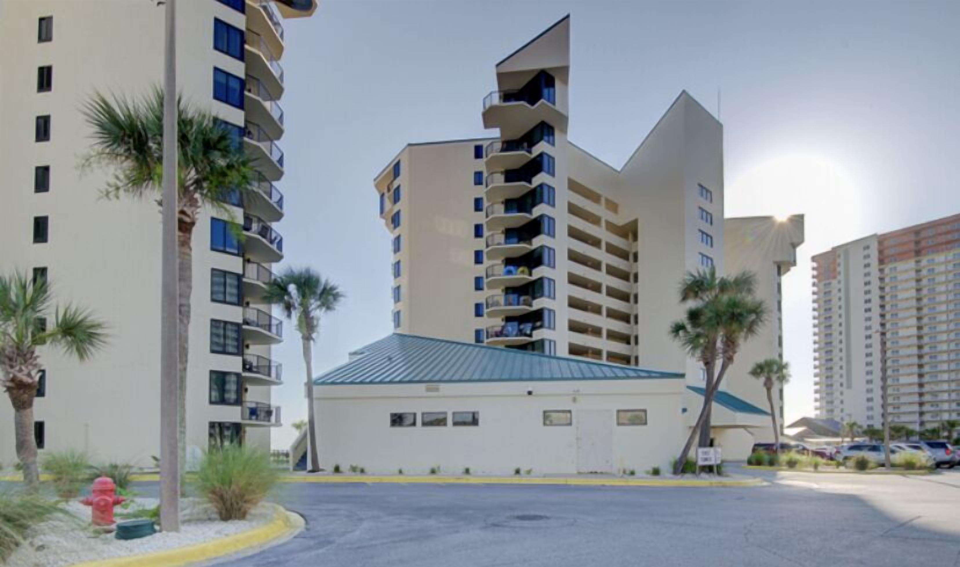 A modern multi-story building with balconies, a parking lot, and palm trees.