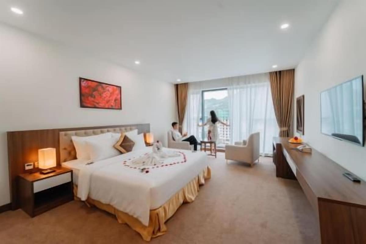 A hotel room with a bed, bedside table, lamp, TV, and a view of the outdoors.