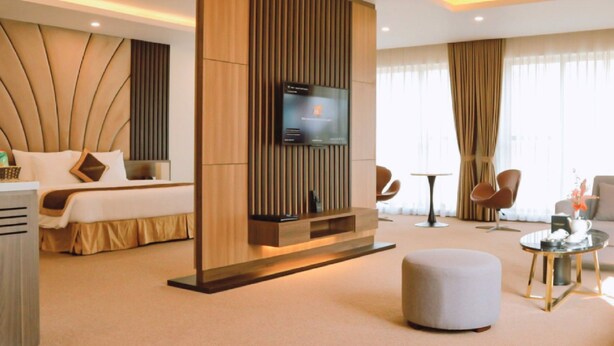 Une chambre d’hôtel moderne avec un lit, un téléviseur fixé sur un panneau en bois, un pouf rond et une petite table avec une lampe.