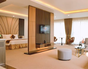 Una habitación de hotel moderna con una cama, un televisor montado en un panel de madera, un otoman redondo y una mesita de noche con una lámpara.