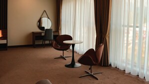 Une chambre d’hôtel équipée d’un bureau, de deux chaises, d’une table ronde, d’un miroir et de grandes fenêtres avec des rideaux.