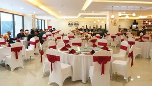 Une salle de banquet avec des tables rondes, des chaises blanches ornées de rubans rouges et un espace scène doté d’un comptoir.