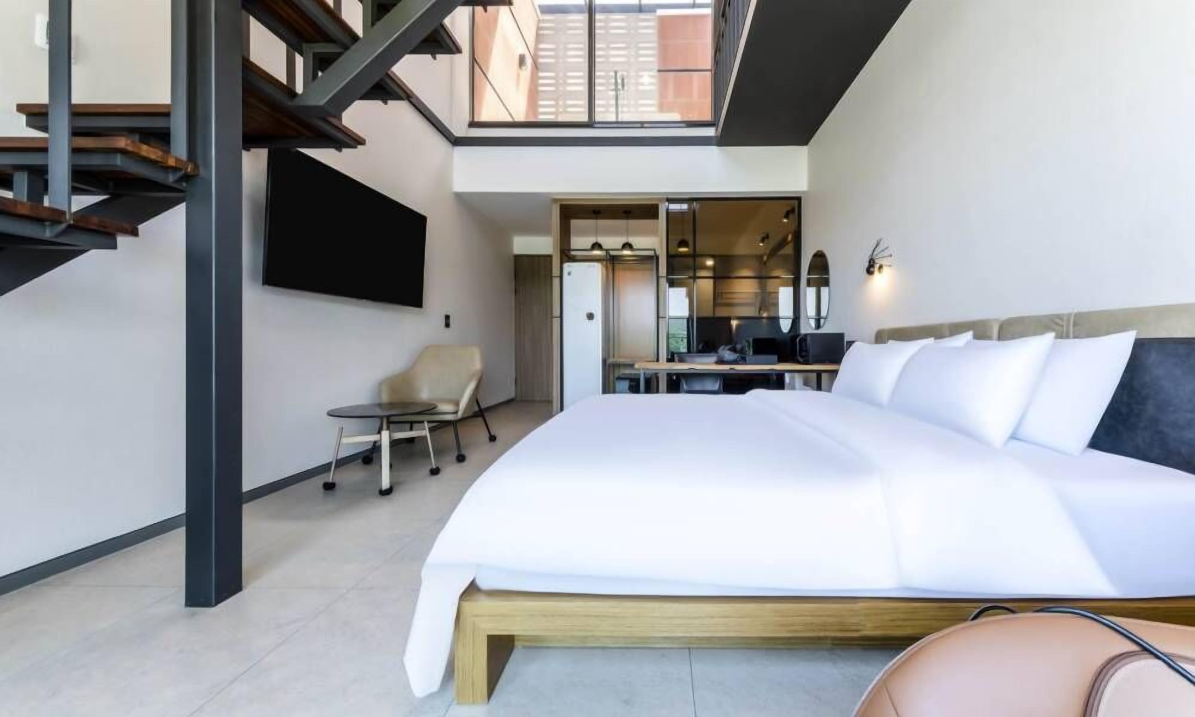 A modern hotel room with a large bed, a small table, a chair, and a kitchen area visible through an open door.