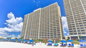 A high-rise hotel with multiple balconies, a beach area with sun loungers and umbrellas, and a clear blue sky.
