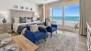 A spacious bedroom with a large bed, two blue armchairs, a wooden coffee table, and a view of the ocean through large windows.