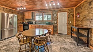 A rustic kitchen with wooden walls, a round wooden table with chairs, a stainless steel refrigerator, a microwave, and a clock on the wall.