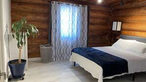 A bedroom with a wooden log wall, a bed with white and dark blue bedding, a potted plant, a waste basket, and a floor lamp.