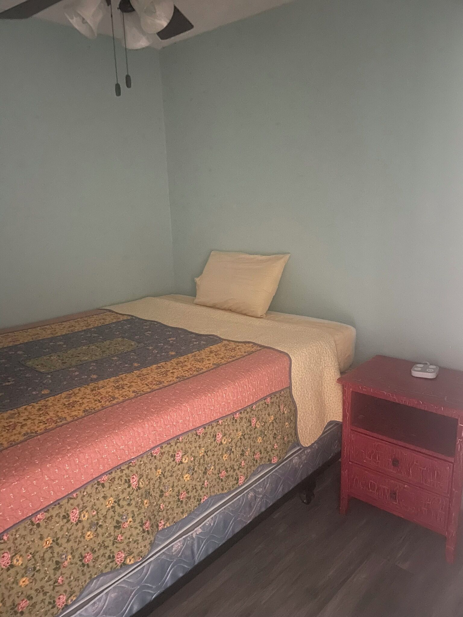 Un lit simple avec une couvre-lit à motifs floraux et un oreiller uni, une petite table de chevet rouge sur laquelle se trouve un objet blanc, et un ventilateur de plafond à deux pales.