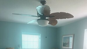 Une chambre à coucher comprenant un lit, des tables de chevet, un ventilateur de plafond, une fenêtre avec des stores et un tableau encadré accroché au mur.