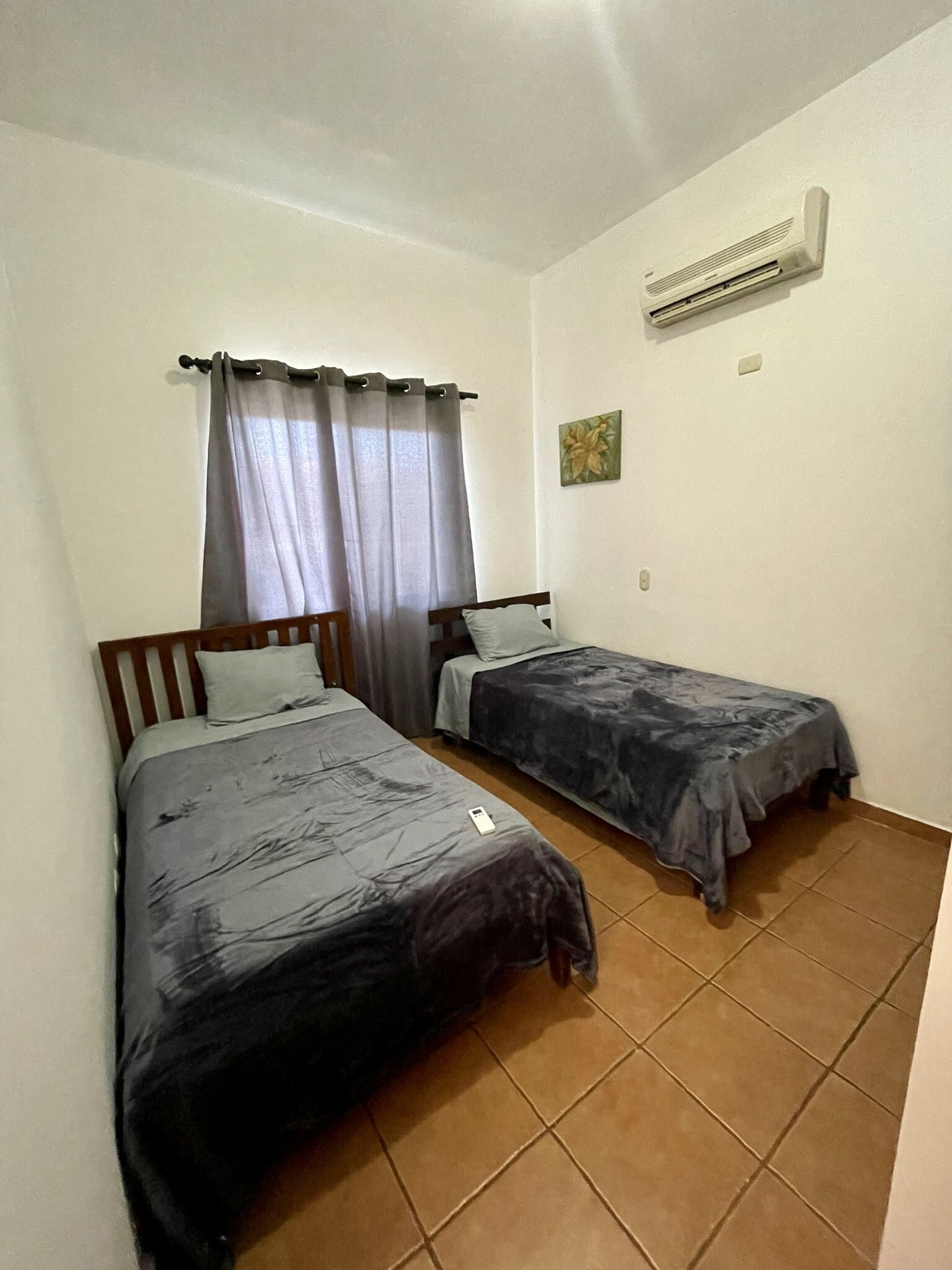 A small, clean room with two single beds, a window with curtains, an air conditioning unit, and a small painting on the wall.