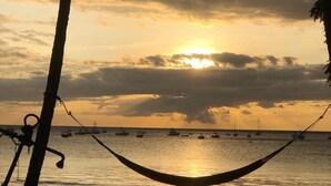 Una hamaca atada entre dos árboles con vista a un mar tranquilo y barcos a lo lejos durante el atardecer.