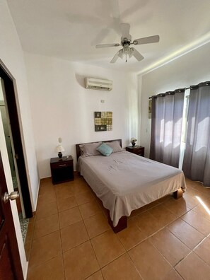 A bedroom with a bed, a ceiling fan, an air conditioner, a nightstand, a lamp, a curtain, and a door. - Beautiful Coco Beach 2 Bedroom Condo (playas del coco)