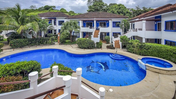 Un hotel con una amplia piscina, varias edificaciones con detalles azules y abundante vegetación.