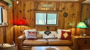 Interior acogedor de una cabaña con un sofá de cuero, una mesa de madera y un cartel que dice 'WELCOME' sobre una ventana.