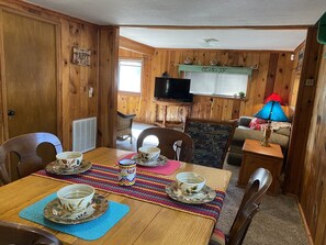 Ein gemütliches Blockhaus-Interieur mit einem Holzesstisch, gedeckt für vier Personen, einem an der Wand montierten Fernseher und einem Kamin mit dekorativem Sims.