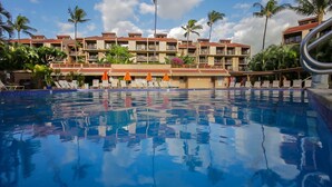 A hotel pool area with multiple lounge chairs and umbrellas, surrounded by palm trees and a multi-story building with balconies. - 🌈Updated OCEANVIEW Kihei Penthouse, Sleeps 6, +Beach Gear🐬 (Kihei)