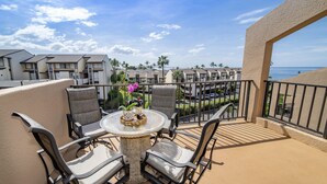 A balcony with outdoor seating, a small table with a decorative item, and a view of a resort area with buildings and palm trees. -   🌈Updated OCEANVIEW Kihei Penthouse, Sleeps 6, +Beach Gear🐬 (Kihei)