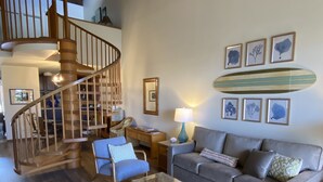 A spacious living area with a wooden staircase, a grey sofa, a coffee table, and a surfboard decoration on the wall. - 🌈Updated OCEANVIEW Kihei Penthouse, Sleeps 6, +Beach Gear🐬 (Kihei)
