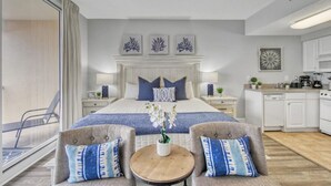 Una habitación de hotel moderna con una cama grande, dos sillones, una mesita, una zona de cocina y vistas a través de un ventanal.