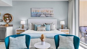 A modern bedroom with a large bed, two bedside tables, a nightstand, a round side table, and a view of the beach through the window. - BeachFront - Ocean View- Majestic Beach Resort 709 (Panama City Beach)