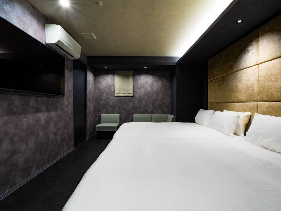 A modern hotel room with a large bed, a flat-screen TV mounted on the wall, a seating area with two chairs, and a small window.
