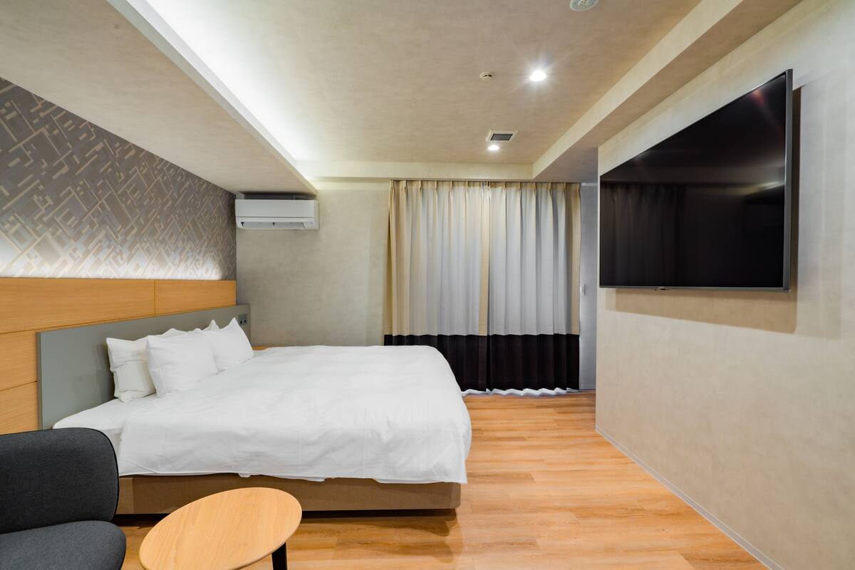 A modern hotel room with a large bed, a flat-screen TV mounted on the wall, and a small wooden table.