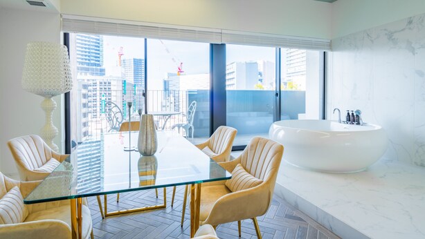 Ett modernt hotellrum med ett glasbord, beige stolar, ett stort fönster och ett vitt badkar.
