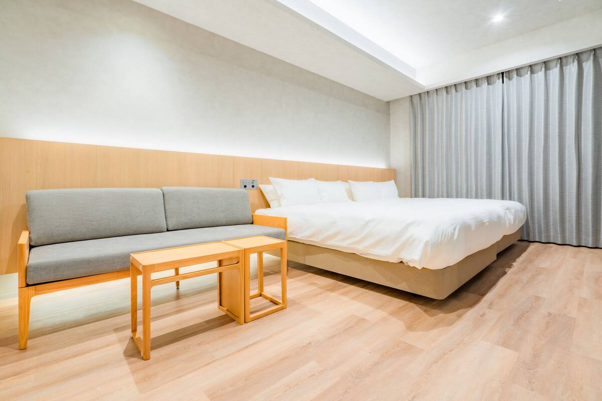 A modern hotel room with a wooden headboard, a grey sofa, a wooden side table, and a large bed with white bedding.