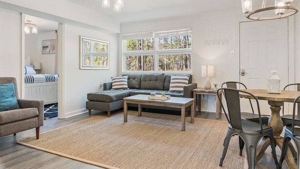 A modern living room with a sofa, coffee table, and dining table. There are large windows with a view of trees.