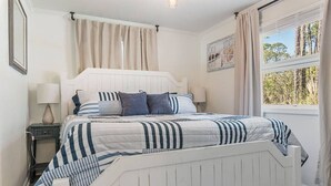 A bedroom with a white bed, striped bedding, a nightstand with a lamp, and a window with curtains.
