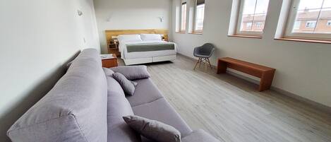Un dormitorio moderno con una cama grande, un sofá gris, un banco de madera y una ventana con vistas a edificios.