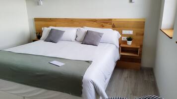 Una cama bien tendida con sábanas blancas y almohadas grises, una mesita de noche con una planta y un cabecero de madera.