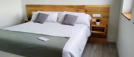 A neatly made bed with white linens and grey pillows, a bedside table with a plant, and a wooden headboard.