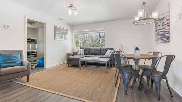 A modern living room with a sofa, coffee table, and dining table with chairs. There is a window providing natural light and a door leading to a kitchen.
