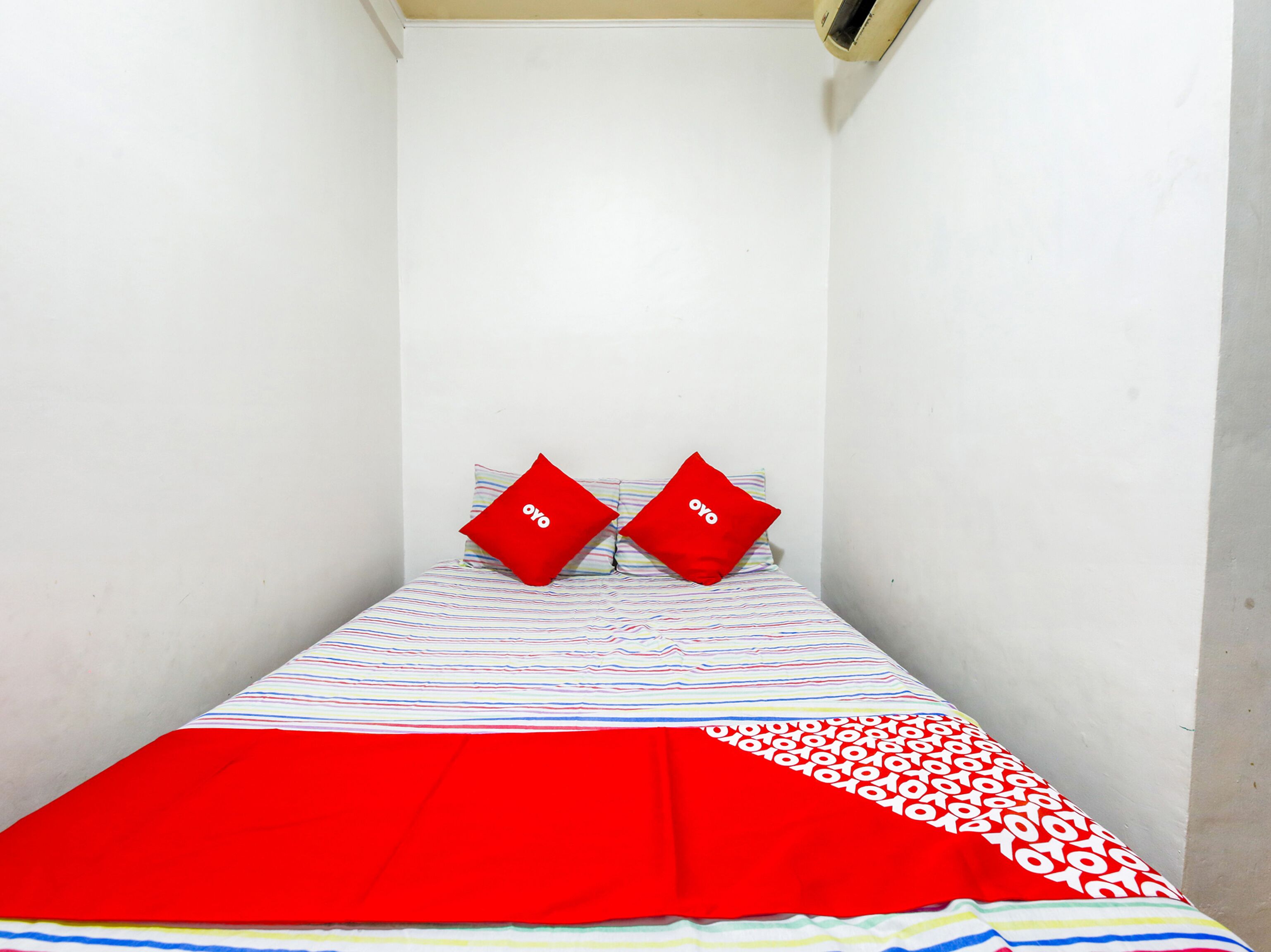 A small, narrow room with a single bed, striped bedding, and two red pillows with the word "oyo" on them.
