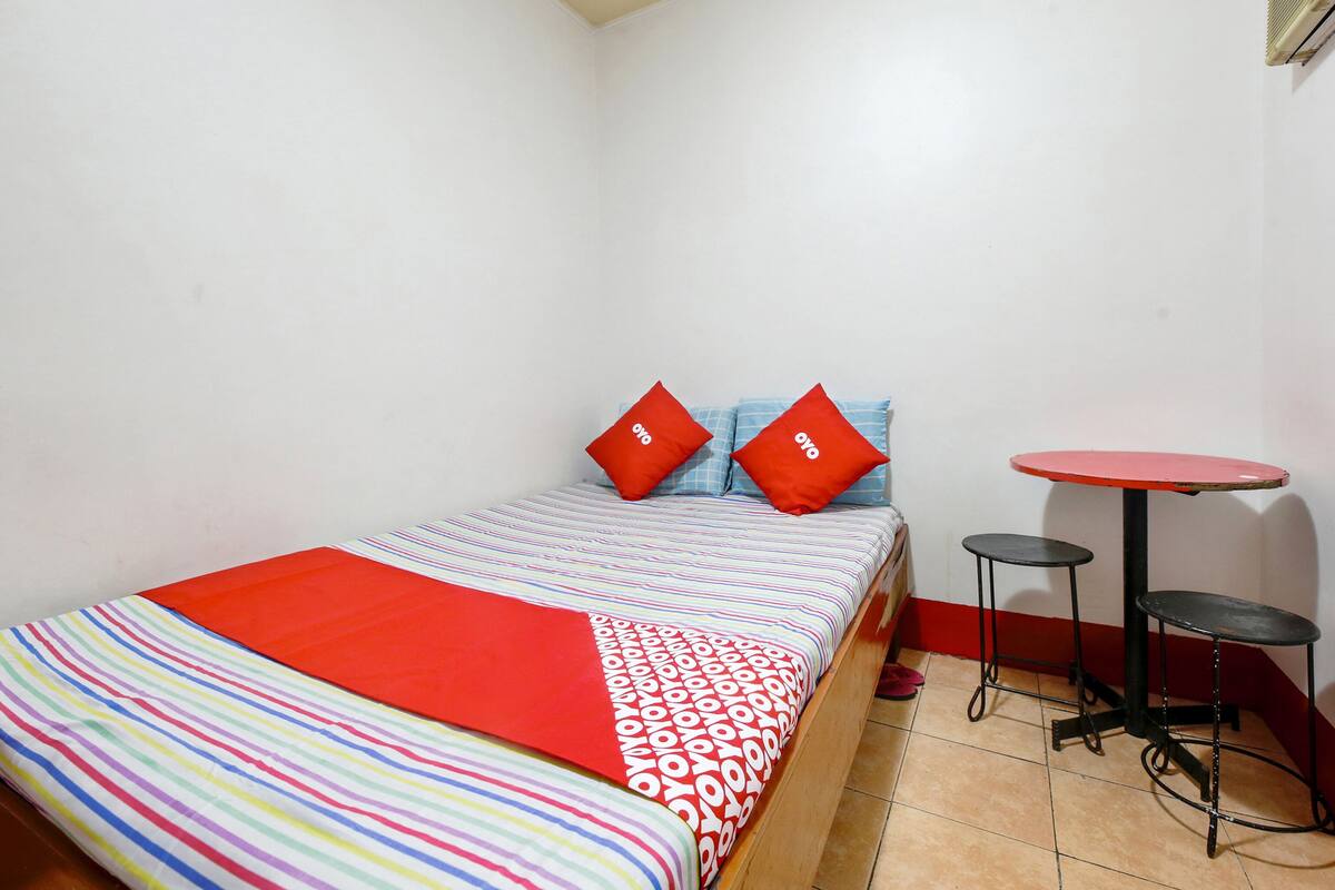A small, neatly arranged room with a bed, two pillows, a striped blanket, and a small round table with a stool.